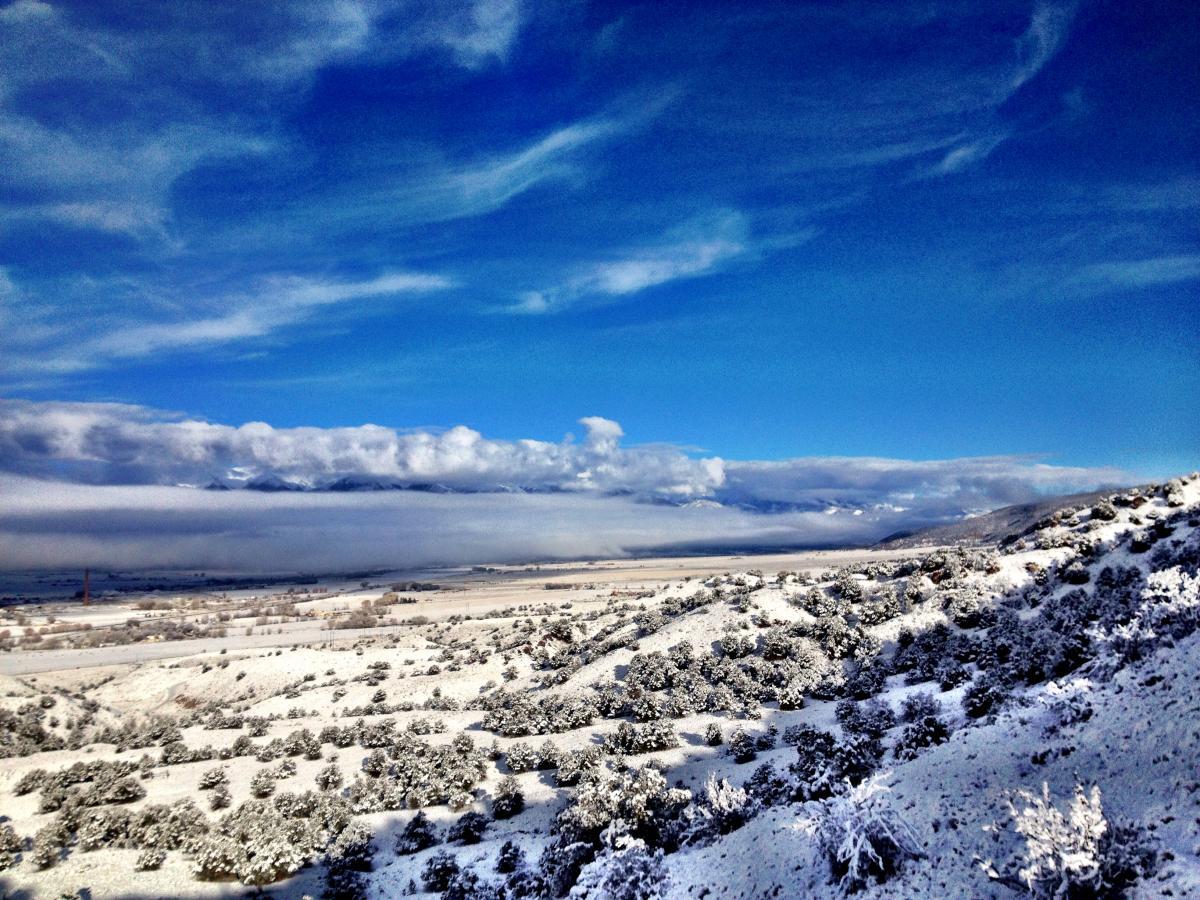 A scenic winter landscape featuring rolling hills blanketed in fresh snow, under a bright blue sky with wispy clouds. In the background, expansive mountains are partially obscured by a layer of fog, creating a tranquil and picturesque view of nature in winter. Arkansas Hills mountain bike trail.