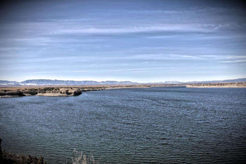 A serene landscape featuring a wide body of water surrounded by rocky shorelines and distant mountains under a clear blue sky. The scene captures the tranquil beauty of nature with rippling water reflecting the sky. South Shore Lake Pueblo mountain bike trail.