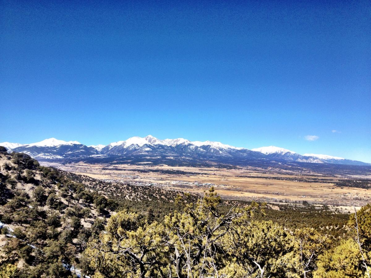 A panoramic view of snow-capped mountains under a clear blue sky, overlooking a valley filled with trees and open fields. The foreground features rocky terrain and greenery, while the distant mountains create a dramatic backdrop. Double Rainbow mountain bike trail.