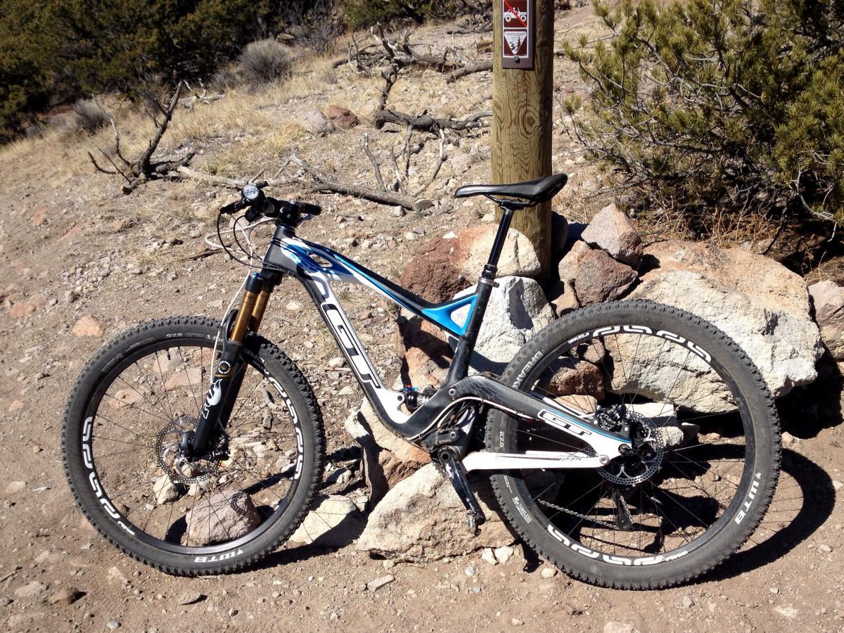GT Force Carbon Pro: A mountain bike parked on rocky terrain, positioned next to a wooden trail sign. The bike features a sleek frame with blue and white accents and black wheels, showcasing its off-road capabilities. The background includes sparse vegetation and a dry, rugged landscape.