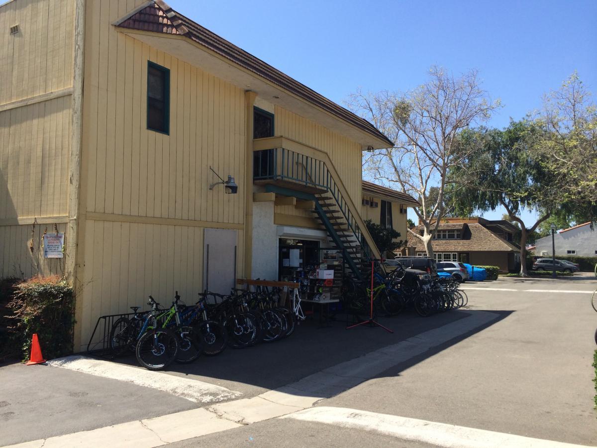 A yellow two-story building with a staircase on the exterior, located in a parking area. Several bicycles are parked in front of the building, with a few leaning against a railing. The scene is set on a sunny day, showcasing clear blue skies and minimal vegetation nearby.