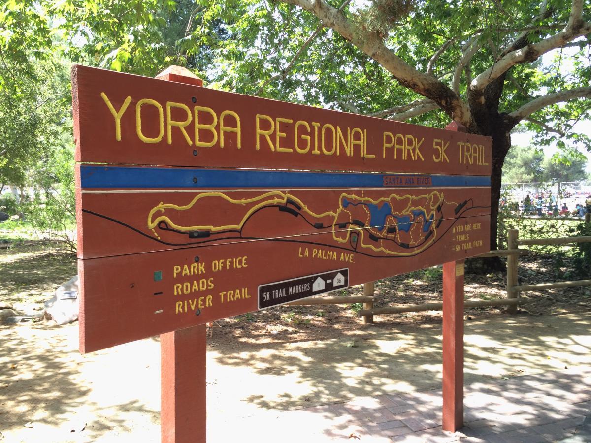 Sign for Yorba Regional Park 5K Trail, featuring a map layout and information about nearby park facilities, including the park office, roads, and river trail. The sign is set against a backdrop of trees, indicating a natural outdoor environment. Yorba Regional Park mountain bike trail.