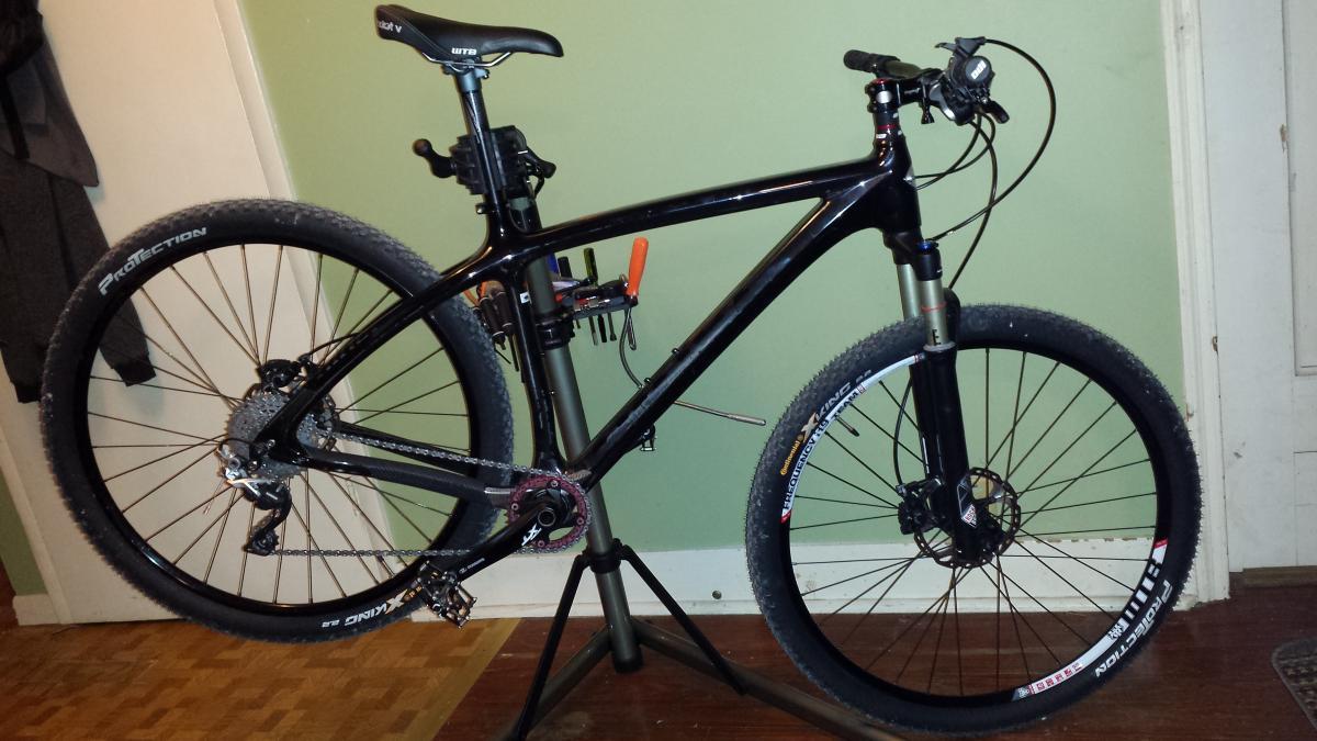 Niner Air 9 Carbon: A black mountain bike displayed on a repair stand, showcasing its components. The bike has thick tires and a sleek frame design. Tools are organized on the repair stand for maintenance. The background features a green wall and a wooden floor.