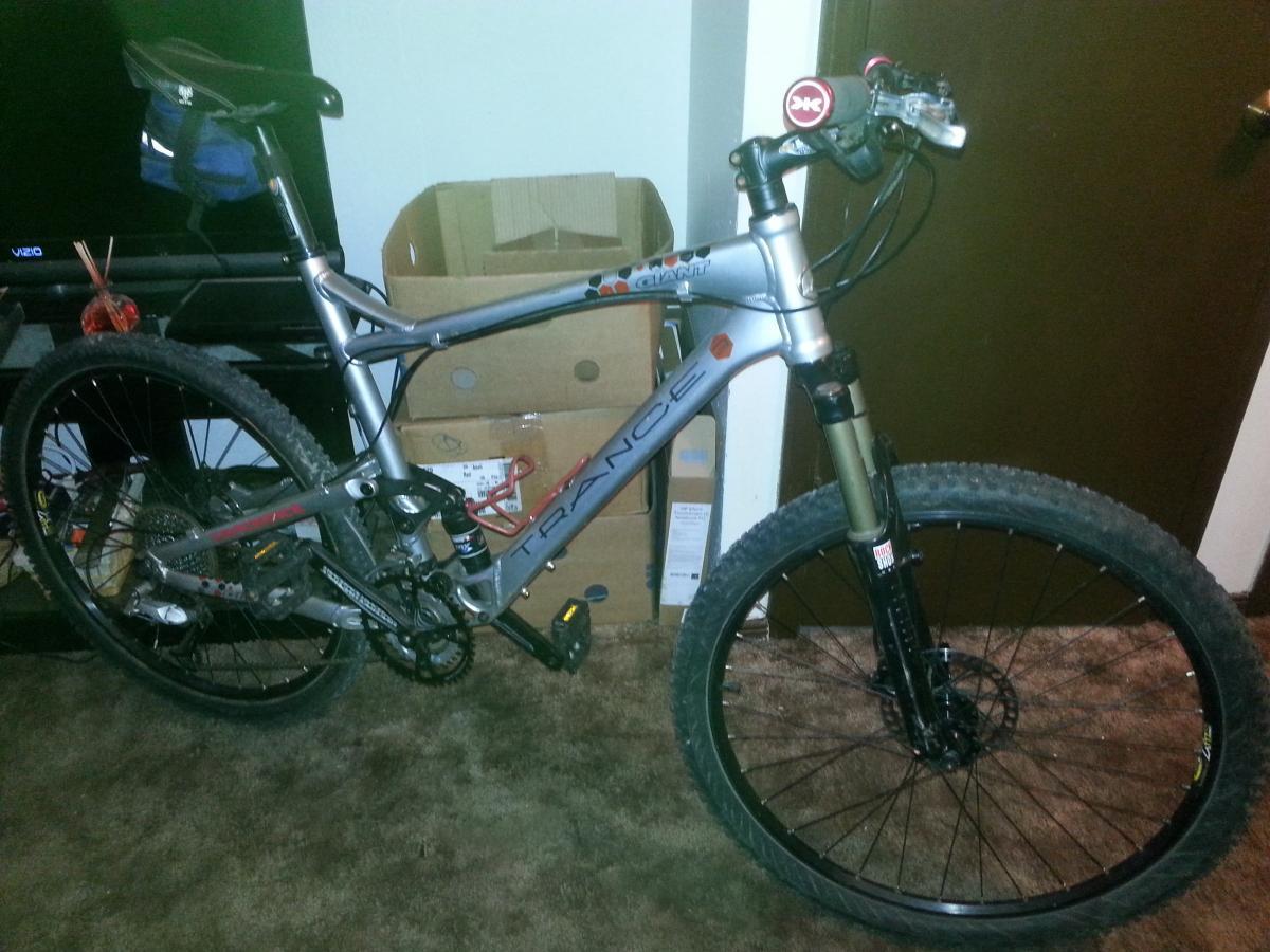 Giant Trance: Alt text: A silver mountain bike leaning against a wall, with a blue pouch attached to the seat and a visible rear wheel. The background features a stack of boxes and a TV. The floor is carpeted.
