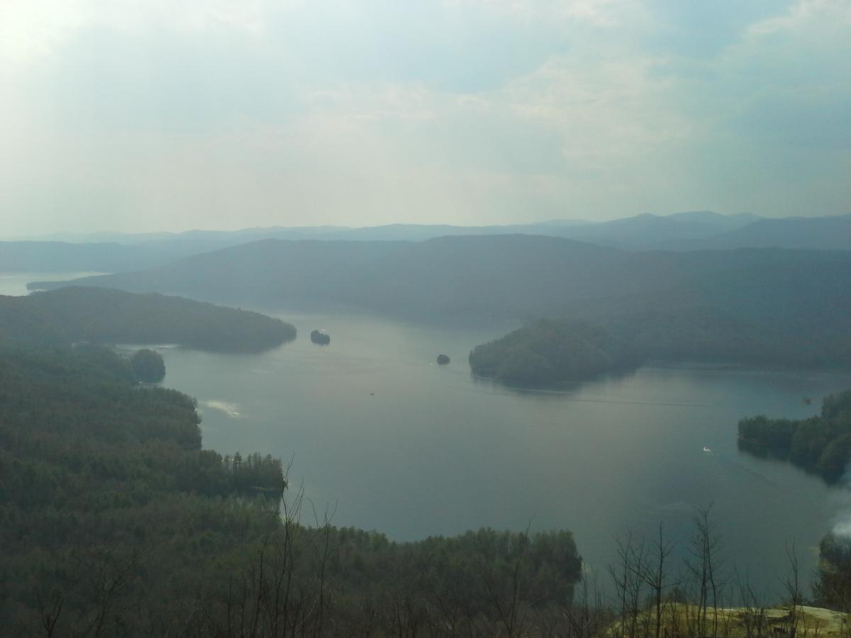 A scenic view of a tranquil lake surrounded by rolling hills and distant mountains under a hazy sky. The water reflects the greenery along the shores and features small islands. A boat can be seen gently moving through the water, enhancing the peaceful atmosphere of this natural landscape. Horse Pasture / Bootleg mountain bike trail.