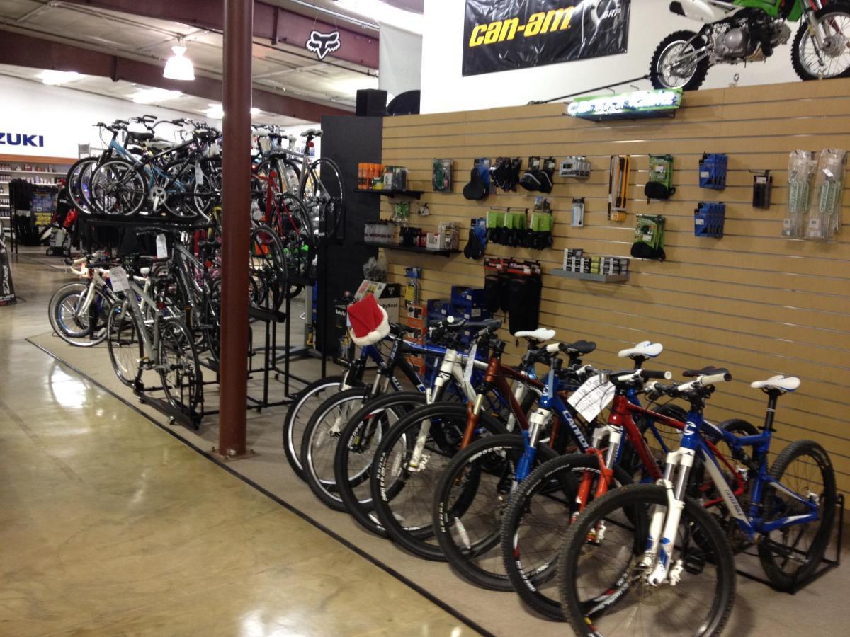 A view of an indoor bike shop displaying various bicycles on racks and shelves. The image features multiple mountain bikes in the foreground and additional bikes hanging on display in the background. Accessories and equipment related to biking can be seen on the walls, along with other merchandise. The shop has a spacious layout with polished concrete floors.