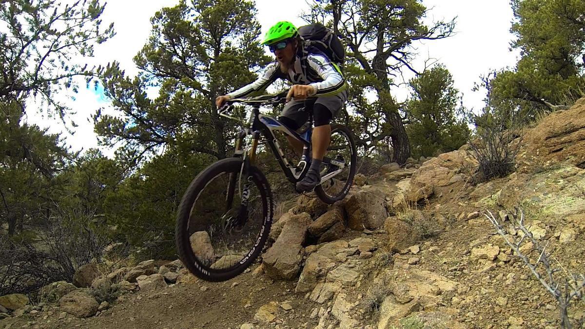A mountain biker in a bright green helmet performs a jump over rocky terrain surrounded by trees. The rider is wearing a white and black jersey and is navigating a challenging trail. Unkle Nazty mountain bike trail.
