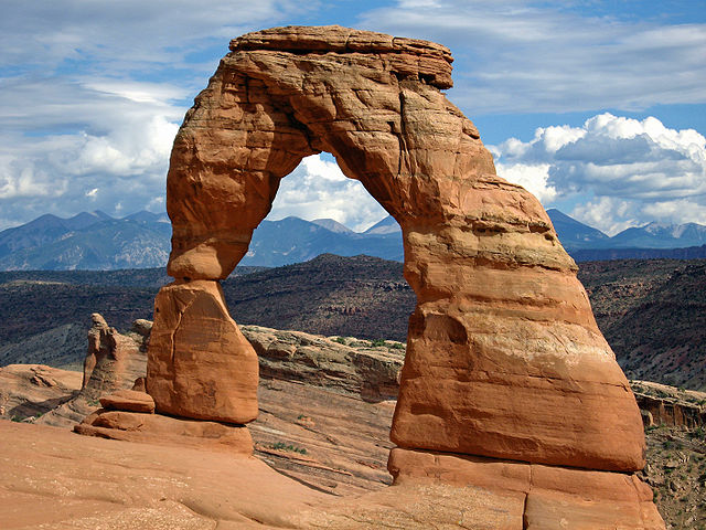 A natural rock formation known as Delicate Arch, characterized by its iconic arch shape, rising prominently from a rocky landscape with distant mountains and a partly cloudy sky in the background.