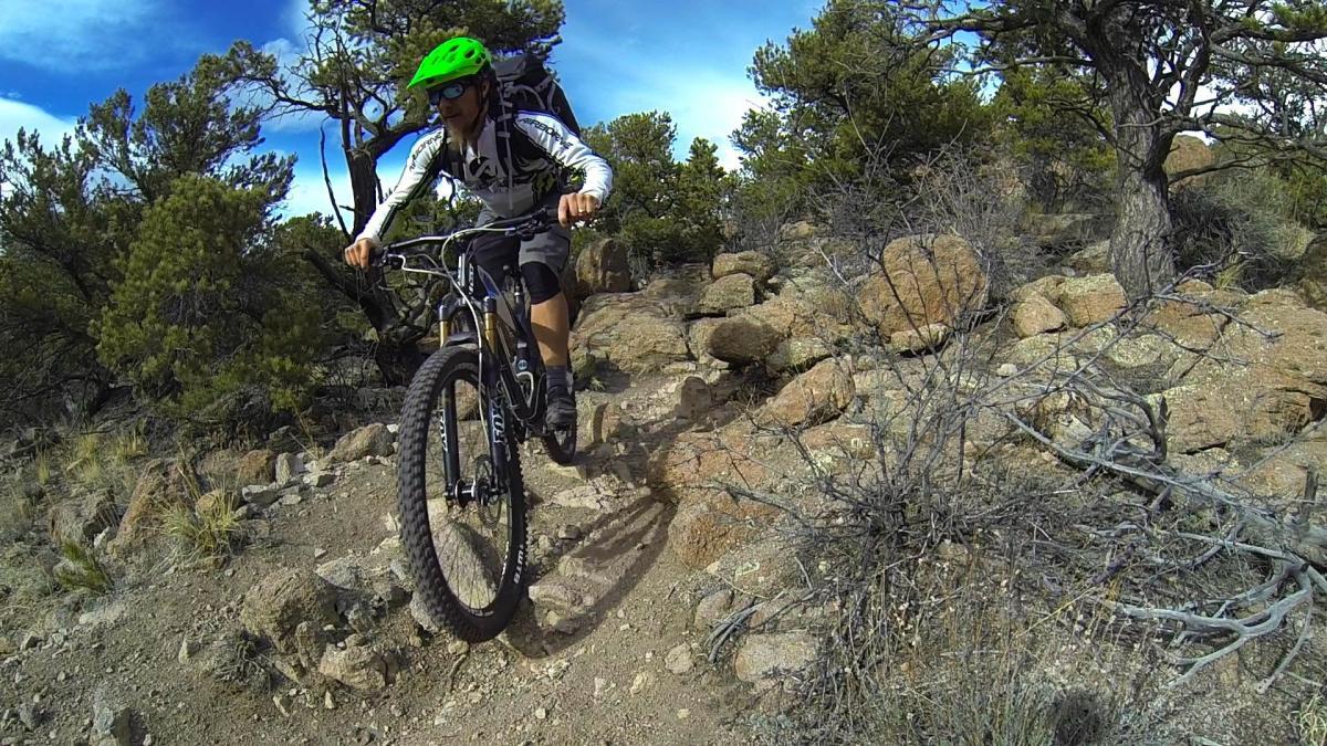 A mountain biker wearing a green helmet and sunglasses navigates over rocky terrain on a trail surrounded by trees and blue skies. Unkle Nazty mountain bike trail.