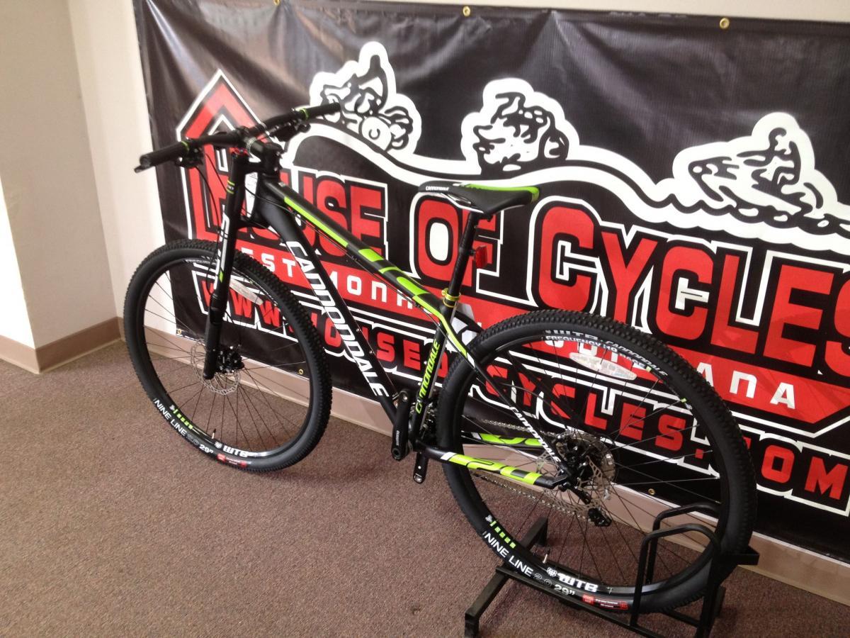 A black and green Cannondale mountain bike displayed on a stand in front of a "House of Cycles" banner, featuring red and white text. The bike has thick tires and a sleek frame, highlighting its sporty design. The floor is carpeted, and the background includes a promotional banner.