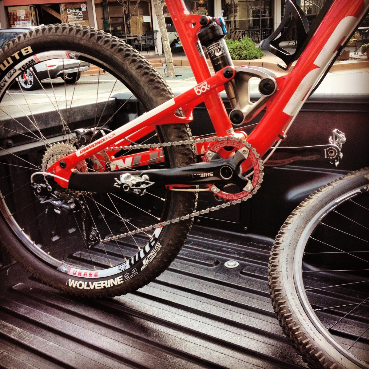 Diamondback Sortie Comp: A close-up view of the rear section of a red and white mountain bike, featuring a Diamondback frame, Shimano crankset, and a WTB Wolverine tire, positioned in the bed of a pickup truck. The bike's chain is clearly visible, along with the suspension components.