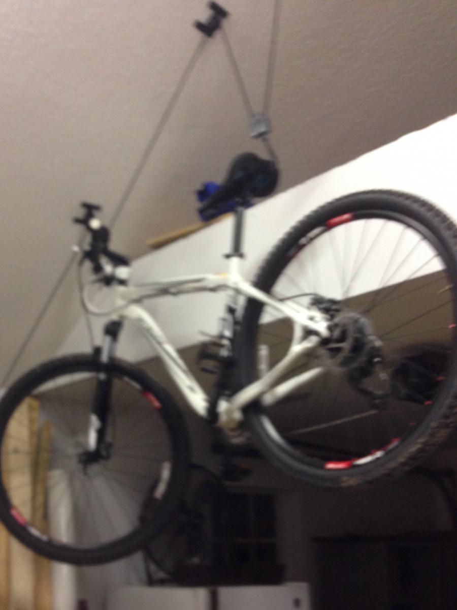 Specialized Rockhopper Comp 29: A white mountain bike hanging from the ceiling in a garage, secured with pulleys and straps, with its wheels and handlebars visible. The background includes wooden boards and a fan.