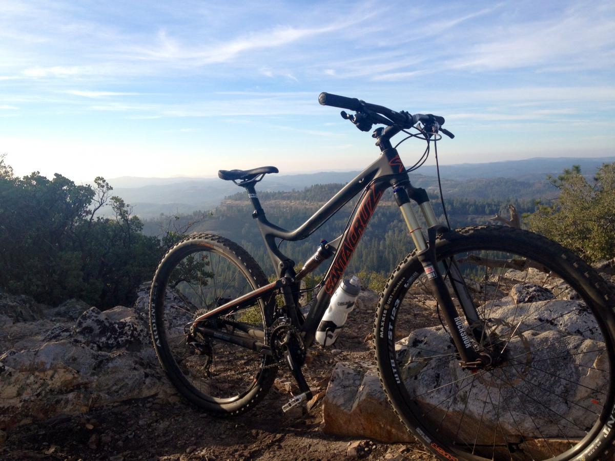 Santa Cruz TallBoyLTc: A mountain bike leaning against a rocky outcrop, set against a picturesque landscape of rolling hills and distant mountains under a clear blue sky. The bike features a black frame with orange accents and is accompanied by a water bottle.