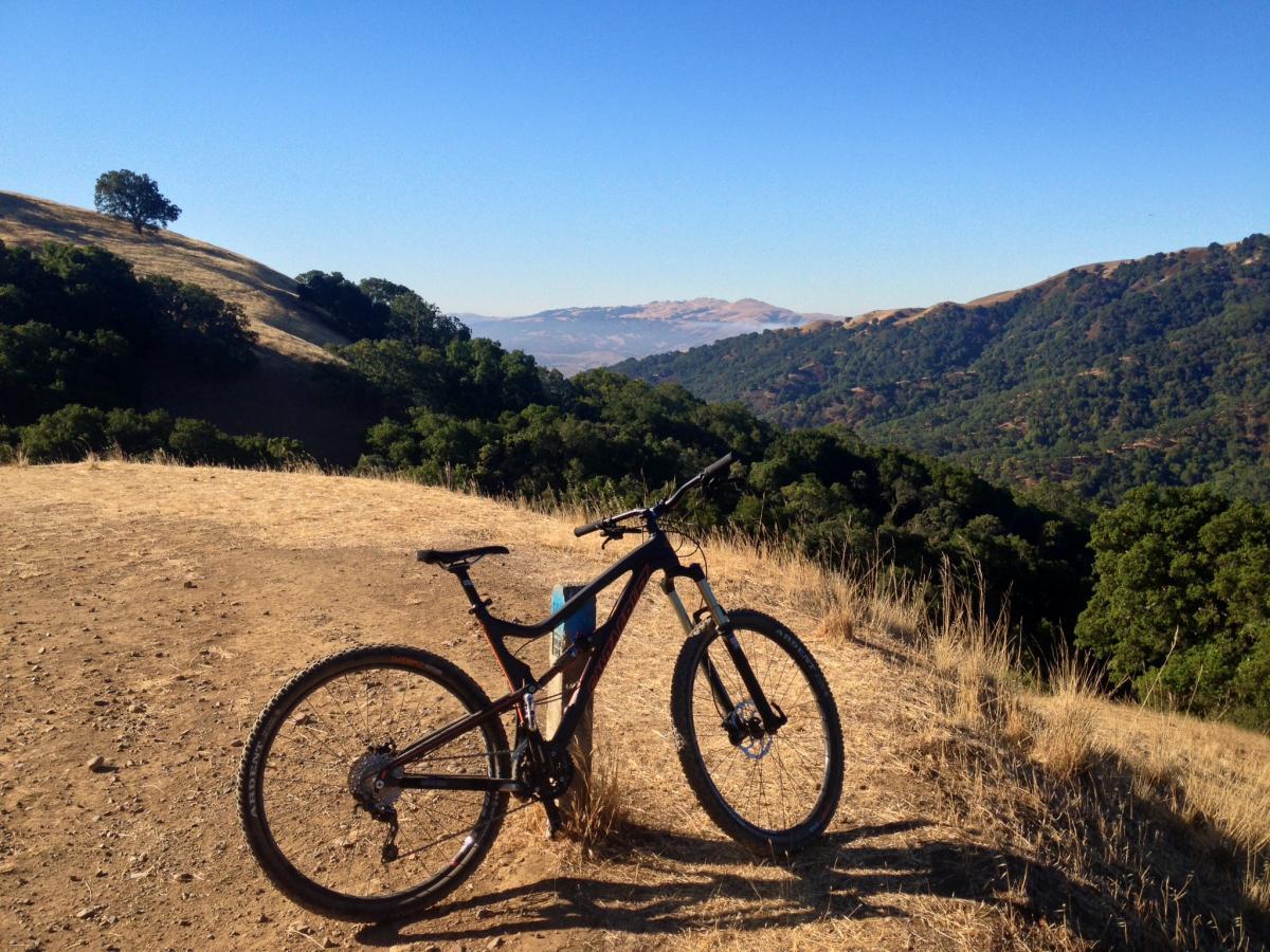 Santa Cruz TallBoyLTc: A mountain bike rests on a dry, dirt path overlooking a scenic valley filled with green trees and rolling hills under a clear blue sky. A lone tree stands on a hillside in the background, enhancing the natural landscape.