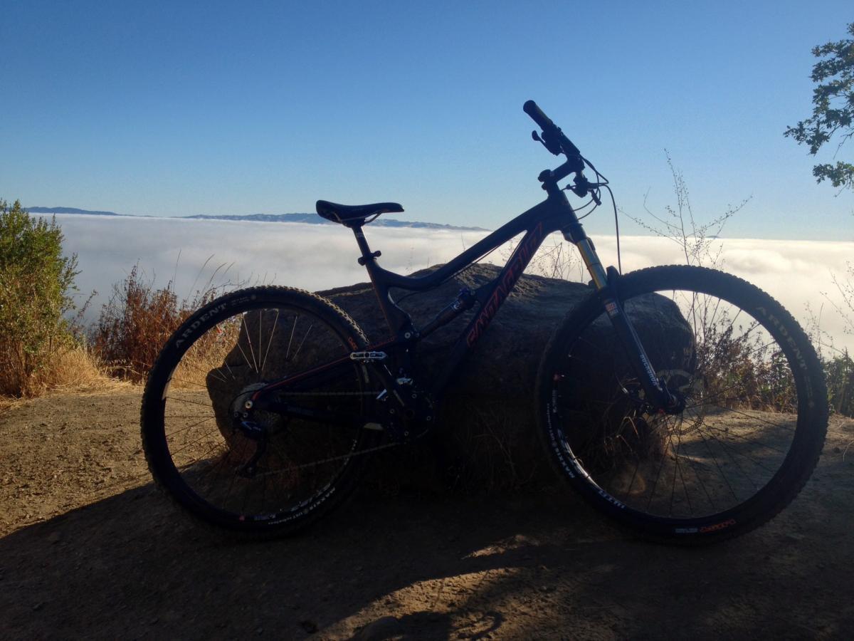 Santa Cruz TallBoyLTc: A mountain bike resting on a rocky surface with a scenic view of clouds below and distant mountains in the background. The sky is clear and blue, emphasizing the elevation of the location. Surrounding vegetation includes dry grass and sparse foliage.