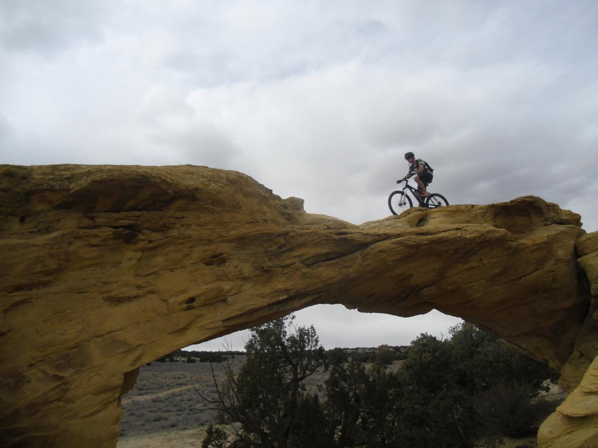 A mountain biker navigating a rocky arch under a cloudy sky, with a desert landscape in the background. Devil
