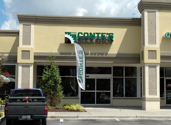 Exterior of Conte's Bike Shop featuring a large sign with the shop's name and the year established. The storefront has glass windows displaying bicycles inside, and an "OPEN" sign is visible. A parked pickup truck is in front of the shop, surrounded by greenery. The sky is partly cloudy.