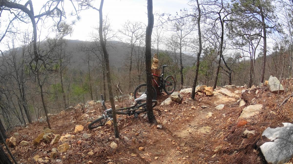 A mountain biking trail featuring two bicycles, one resting against a tree and the other lying on the ground, surrounded by rocky terrain and sparse trees in a hilly landscape. The atmosphere is serene with a cloudy sky and distant mountains visible in the background. Coldwater Mountain mountain bike trail.