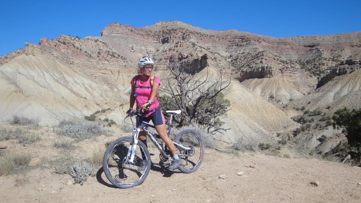 A woman in a pink cycling outfit stands next to her mountain bike on a dirt trail, surrounded by rugged, colorful rock formations under a blue sky. 18 Road Trails / North Fruita Desert mountain bike trail.
