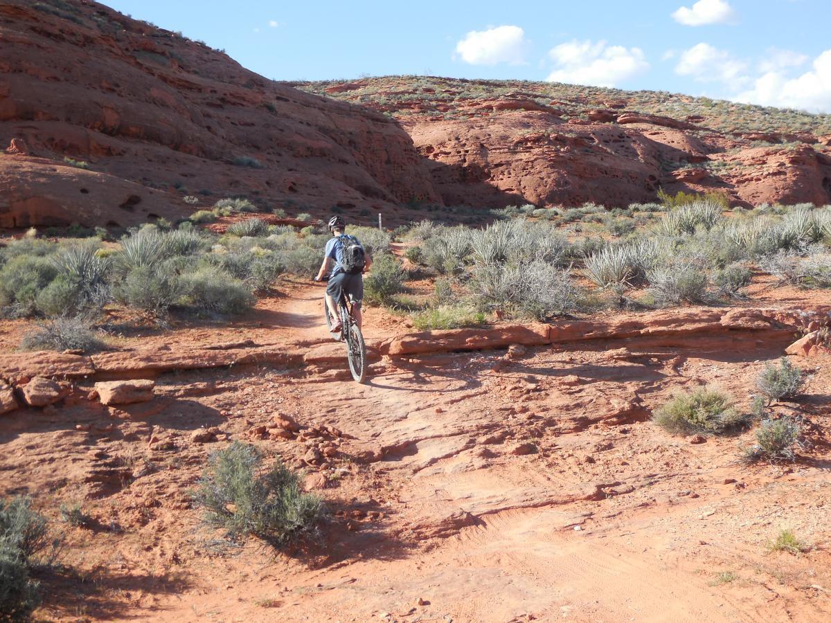 A cyclist riding a mountain bike along a rocky trail in a desert landscape, surrounded by reddish terrain and sparse vegetation under a partly cloudy sky. Prospector - Church Rocks Loop mountain bike trail.