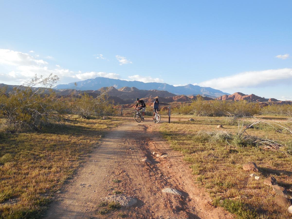 Two cyclists pause on a dirt path surrounded by desert vegetation, with rugged mountains in the background under a blue sky. The landscape is characterized by dry brush and rocky terrain, suggesting an outdoor adventure setting. Prospector - Church Rocks Loop mountain bike trail.