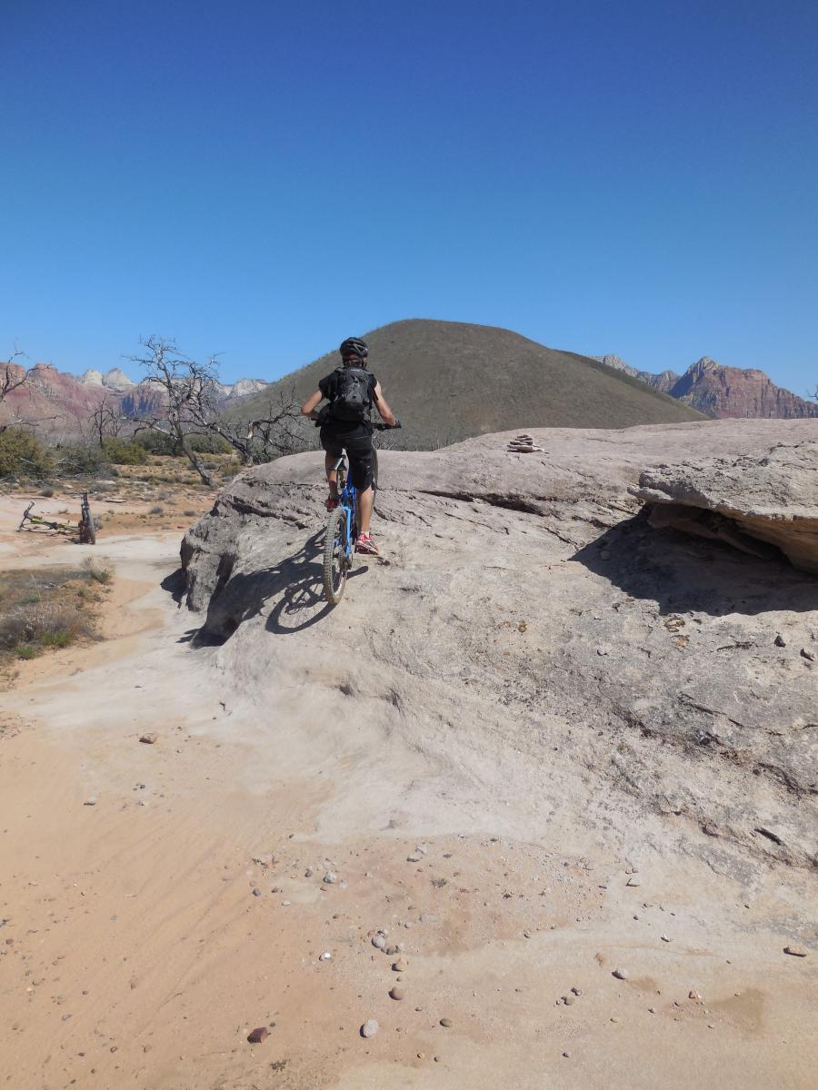 A person riding a mountain bike along rocky terrain with a clear blue sky above and desert landscape in the background. The cyclist is focused on navigating the rocky surface, while hills and distant mountains are visible in the distance. Guacamole Mesa mountain bike trail.