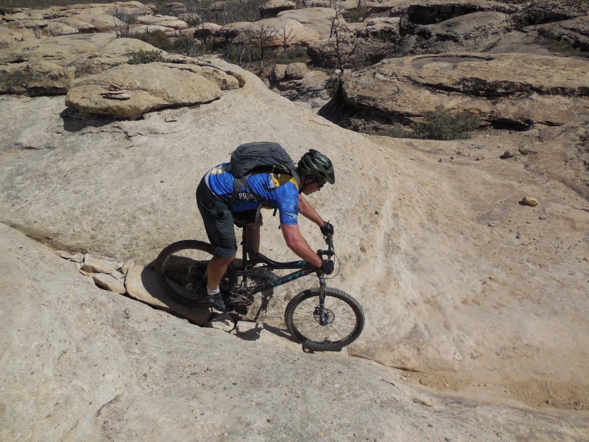 A mountain biker riding on a rocky trail, navigating a steep incline with a rugged landscape in the background. The cyclist is wearing a helmet and a blue jersey, focused on maintaining balance while maneuvering over the uneven terrain. Guacamole Mesa mountain bike trail.