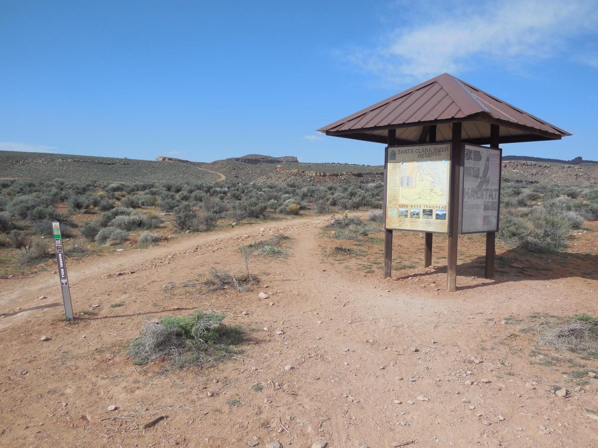 A dirt trail forks at the entrance to the Santa Clara River Reserve, marked by a wooden information kiosk displaying a map and local habitat information. Surrounding the area are sparse shrubs and a rocky terrain under a clear blue sky. Suicidal Tendencies mountain bike trail.