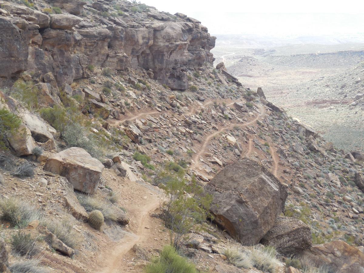 A narrow dirt trail winding through rocky terrain, flanked by steep cliffs and scattered boulders. The landscape features sparse vegetation and a distant view of rolling hills under an overcast sky. Suicidal Tendencies mountain bike trail.
