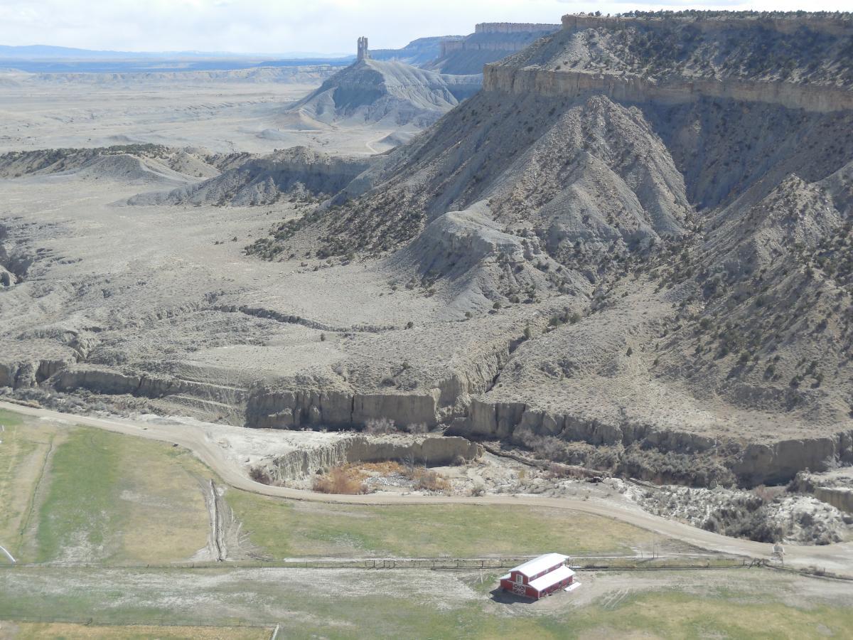 A panoramic view of a rugged landscape featuring steep, gray hills and cliffs, with a distant, tall structure on a plateau. In the foreground, there is a green field with a red barn. The sky is partly cloudy, and the terrain appears arid, suggesting a remote and barren environment. Country Club Trails mountain bike trail.