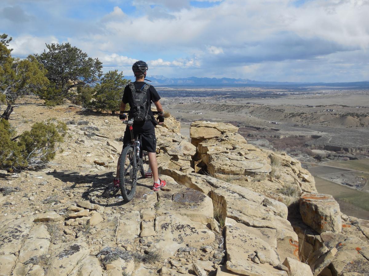 A person standing on a rocky outcrop, overlooking a vast valley with mountain ranges in the distance. They are wearing a helmet and a backpack, and are positioned next to a mountain bike. The sky is partly cloudy, and the landscape features rocky terrain and sparse vegetation. Country Club Trails mountain bike trail.