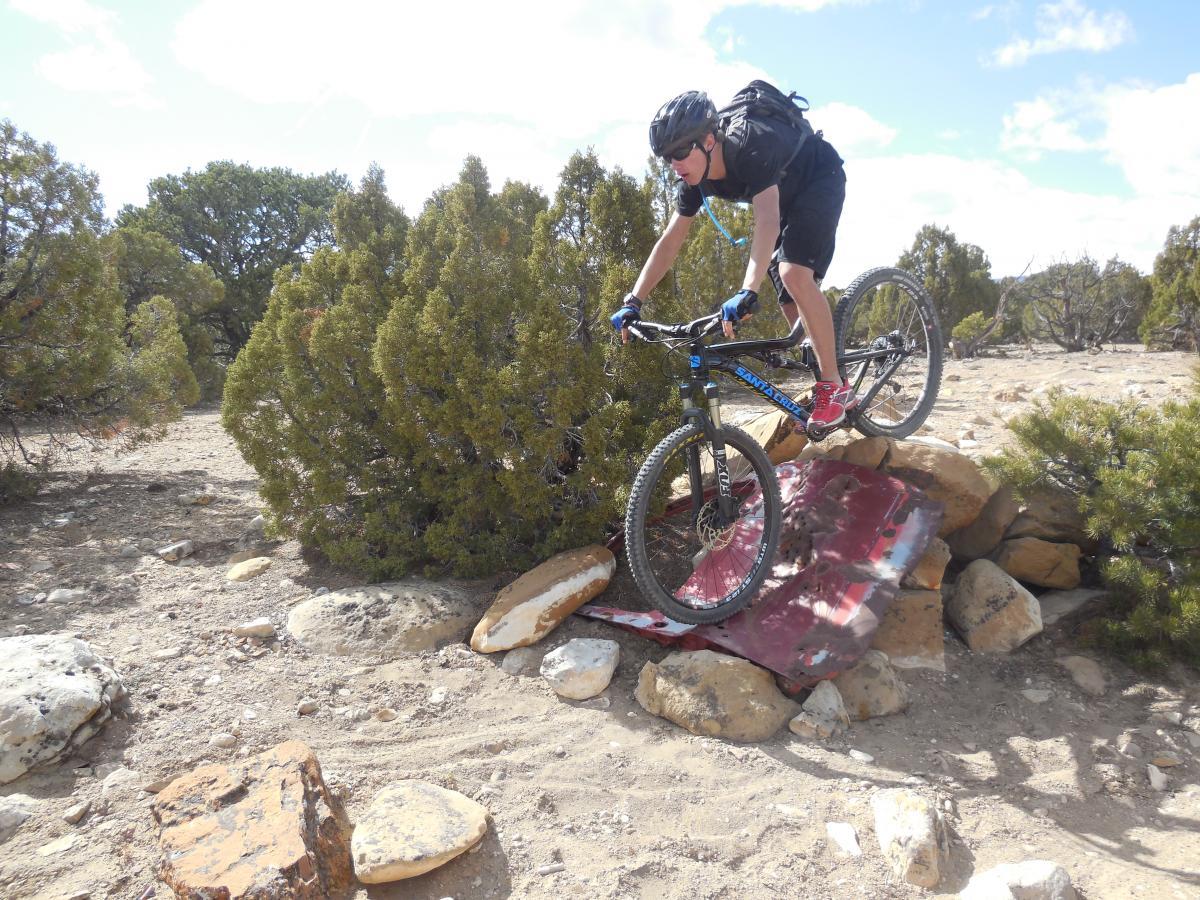 A mountain biker performs a stunt by jumping over a rocky obstacle on a dirt trail, surrounded by shrubs and trees under a partly cloudy sky. Wyatt