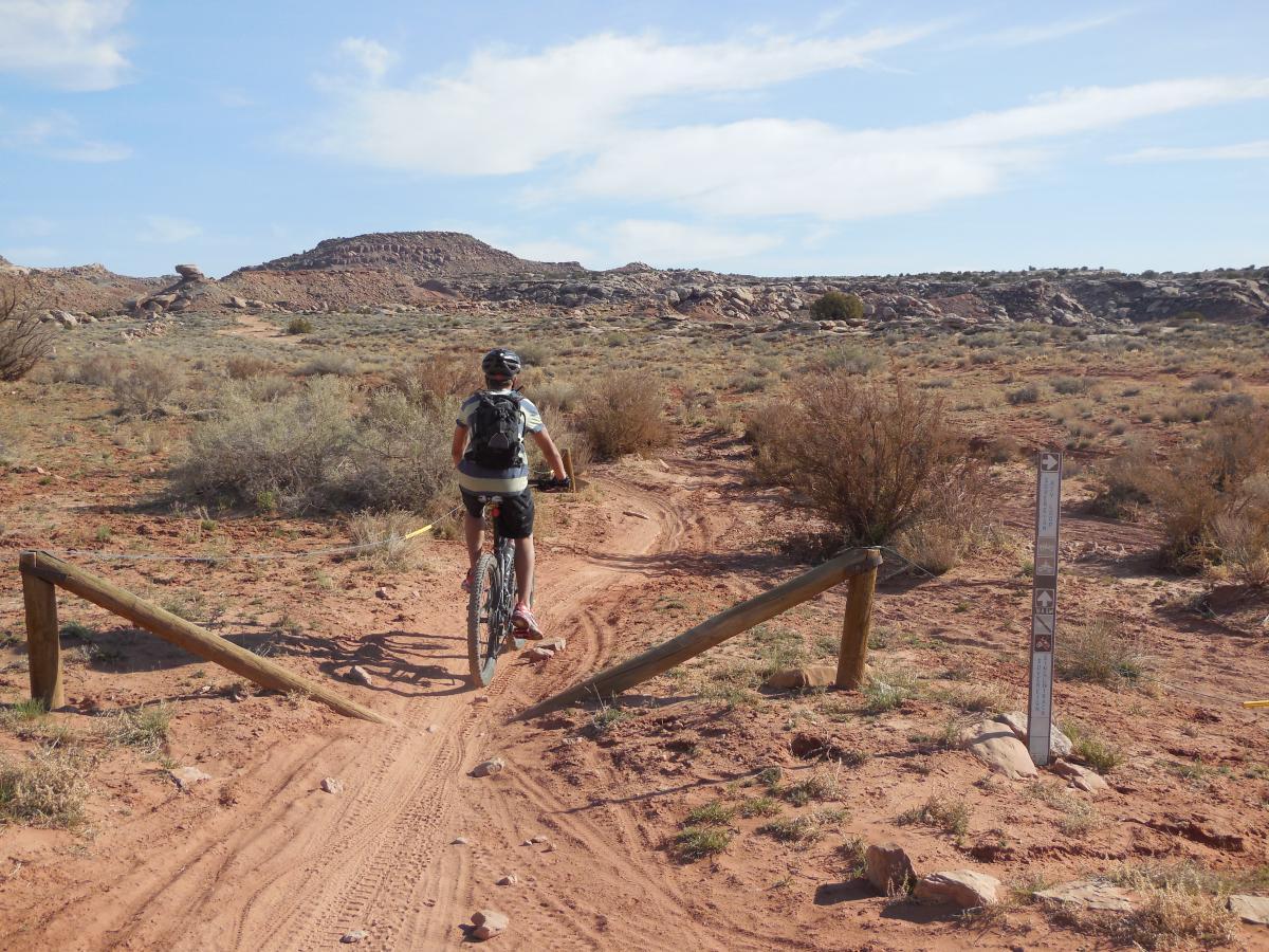 A cyclist rides on a dirt trail surrounded by shrubs and sparse vegetation, leading towards a rocky hillside under a partly cloudy sky. A trail sign stands nearby, indicating path directions. Sovereign Single Track mountain bike trail.