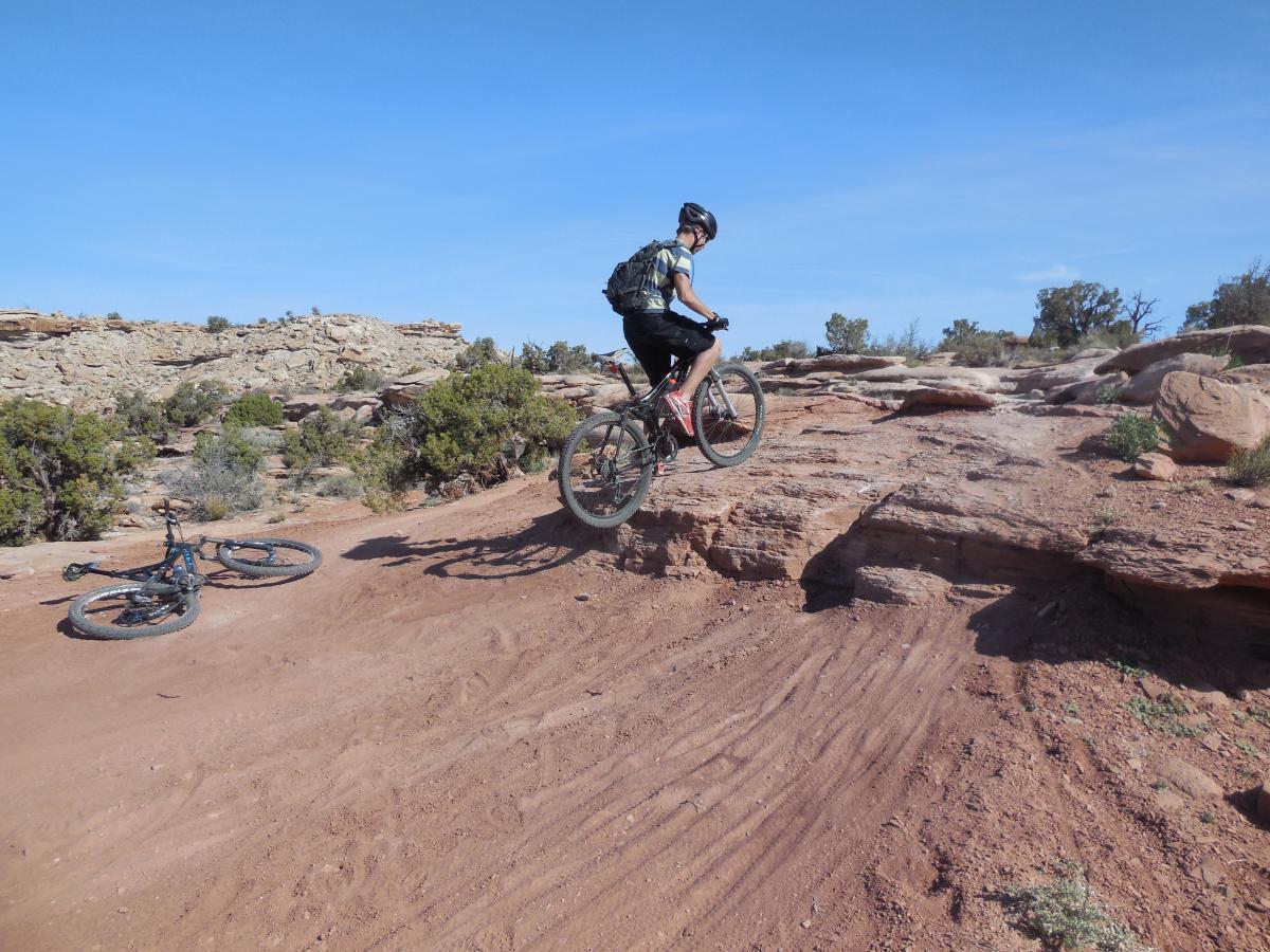 A mountain biker performing a jump over a rocky surface in a desert landscape. Two bicycles are visible on the ground nearby, surrounded by sparse vegetation and rocky terrain under a clear blue sky. Sovereign Single Track mountain bike trail.