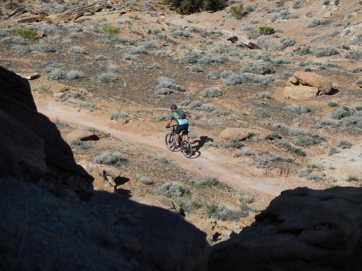A person riding a mountain bike on a dirt trail surrounded by sparse vegetation and rocky terrain. The scene captures the rugged beauty of the outdoors with various shrubs and rocky outcrops in the background. Sovereign Single Track mountain bike trail.