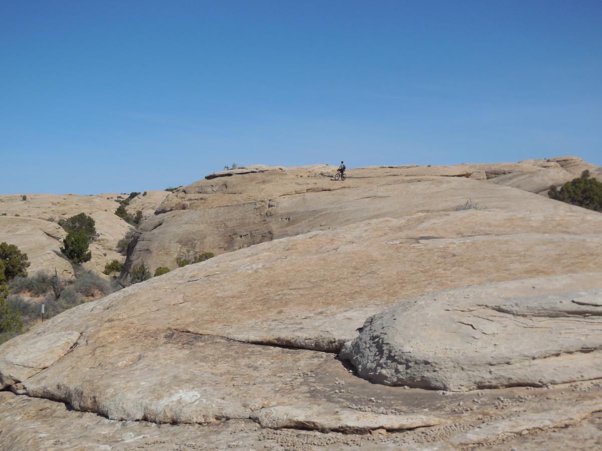 A rocky landscape with smooth, weathered stones under a clear blue sky. In the distance, a person riding a bicycle can be seen atop the rock formation, surrounded by sparse vegetation and hills. The scene captures the natural beauty of the area. Sovereign Single Track mountain bike trail.