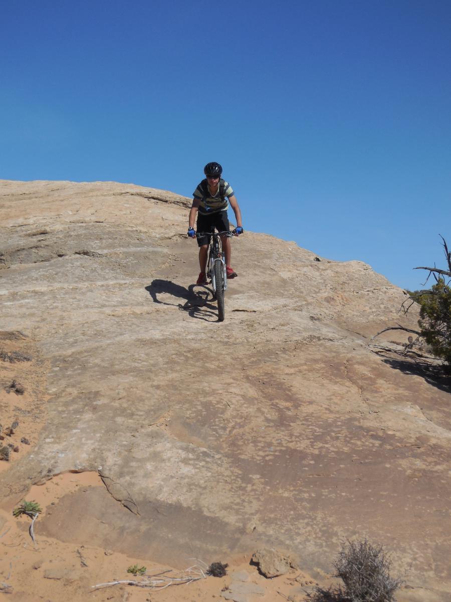A person riding a mountain bike on a rocky terrain under a clear blue sky. The cyclist is positioned on a steep incline, showcasing the challenges of the trail. Sparse vegetation surrounds the area, emphasizing the rugged landscape. Sovereign Single Track mountain bike trail.