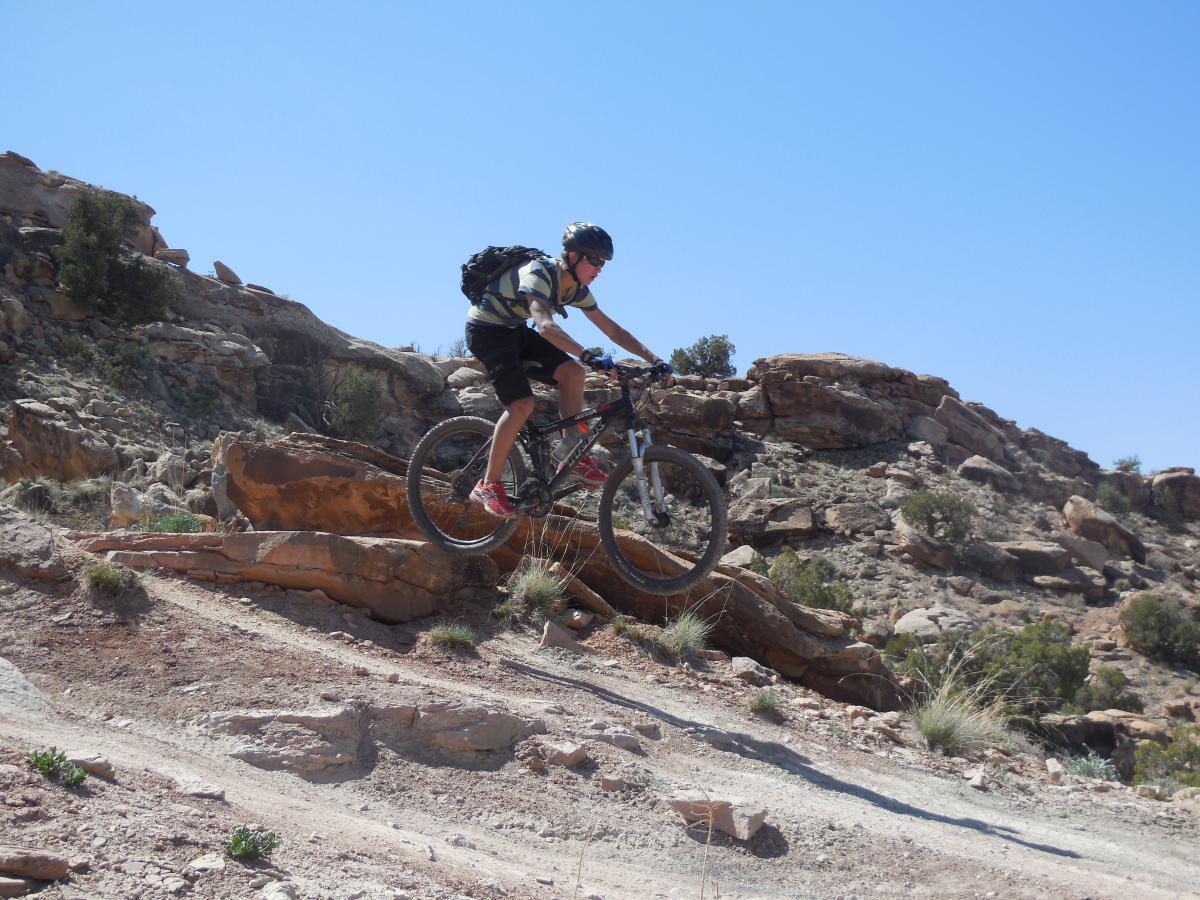 A person riding a mountain bike over rocky terrain, with a clear blue sky in the background. The cyclist appears to be mid-jump, navigating a rocky outcrop in a mountainous area. Sovereign Single Track mountain bike trail.