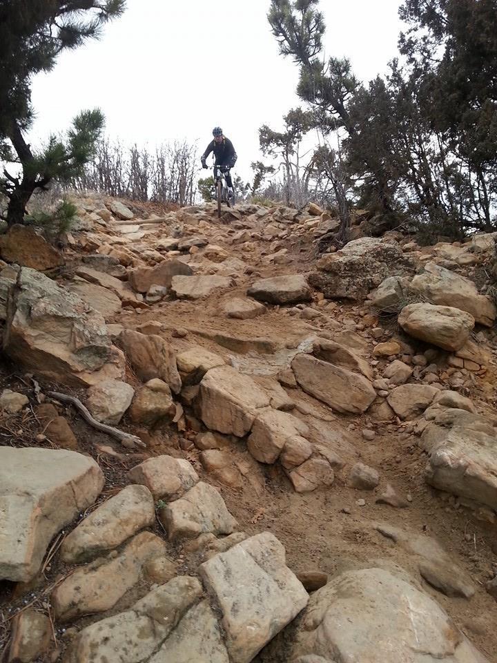 A mountain biker navigating a rocky, uneven trail surrounded by sparse vegetation and trees. The terrain is steep and comprised of various sizes of stones and dirt, suggesting a challenging riding experience. The sky is overcast, indicating possibly cool or damp weather conditions. Red Rocks / Dakota Ridge mountain bike trail.