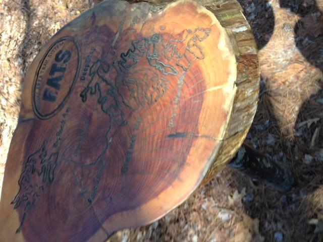 Close-up view of a wooden slab with engraved text and a map design on its surface, set against a natural background of leaves and pine needles. Forks Area Trail System (FATS) mountain bike trail.