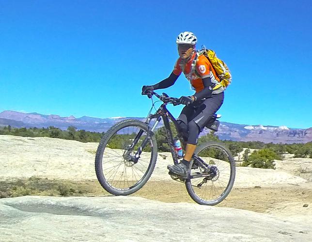 A mountain biker performing a stunts on rocky terrain, wearing a bright orange shirt, black pants, and a helmet. A backpack is secured on the rider's back, and the background features a clear blue sky and distant mountains. Gooseberry Mesa mountain bike trail.