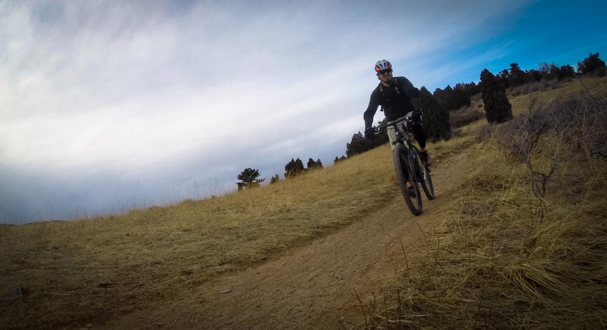 A person riding a mountain bike along a dirt trail in an open grassy area, with a cloudy sky in the background and trees in the distance. Red Rocks / Dakota Ridge mountain bike trail.