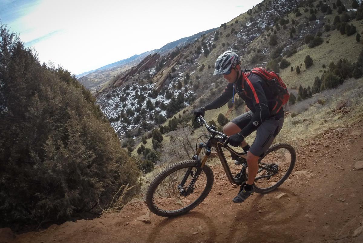 A mountain biker navigates a rugged trail, surrounded by rolling hills and sparse vegetation. The biker wears a helmet and red backpack while riding on a dirt path, showcasing a dynamic outdoor scene with a scenic landscape in the background. Red Rocks / Dakota Ridge mountain bike trail.