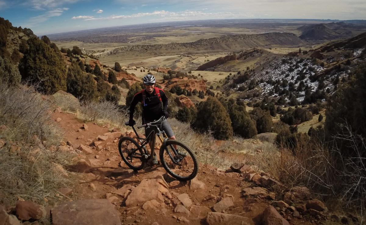 A mountain biker navigates a rocky trail, standing next to his bike on a hillside. The landscape features rolling hills, scattered trees, and a vast view of the valley below under a partly cloudy sky. Red Rocks / Dakota Ridge mountain bike trail.