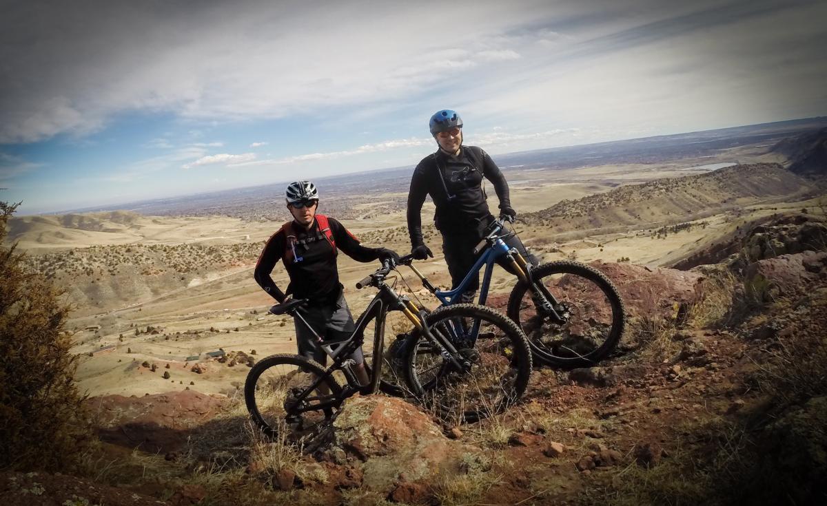 Two mountain bikers pose on a rocky outcrop overlooking a vast landscape of hills and valleys under a partly cloudy sky. They are dressed in cycling gear and helmets, standing next to their mountain bikes, with one person smiling and the other looking at the camera. Red Rocks / Dakota Ridge mountain bike trail.