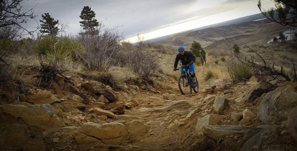 Mountain biker navigating a rocky trail on a hillside, surrounded by sparse vegetation and a cloudy sky. Another cyclist is visible in the background, and the landscape features rolling hills and distant terrain. Apex Park mountain bike trail.