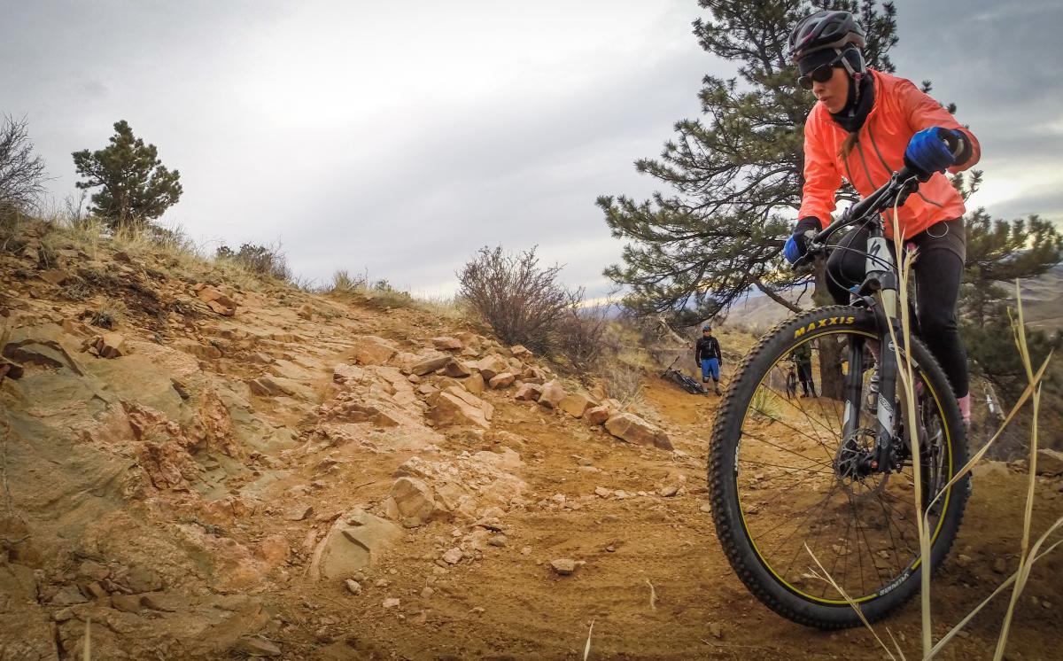 A mountain biker in an orange jacket navigates a rocky path surrounded by sparse vegetation and trees, with another person in the background observing. The sky is overcast, creating a dramatic atmosphere for the outdoor scene. Apex Park mountain bike trail.