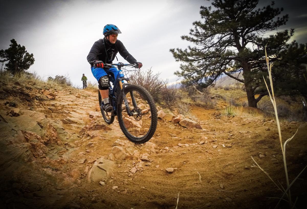 A mountain biker navigating a rocky trail, wearing a blue helmet and protective gear, with a second cyclist visible in the background. The landscape features sparse vegetation and rocky terrain under an overcast sky. Apex Park mountain bike trail.