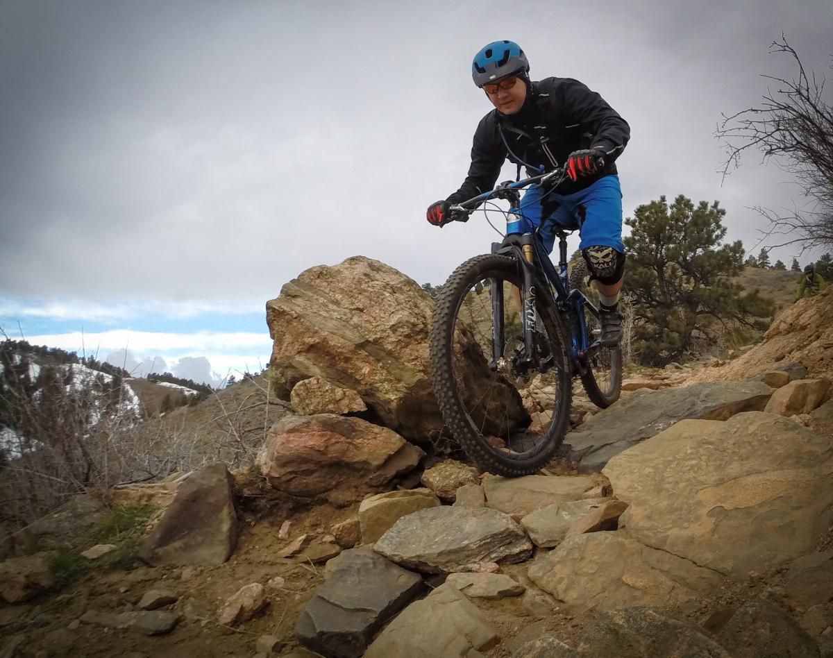 A mountain biker navigates a rocky trail, featuring a mix of large boulders and uneven terrain, under a cloudy sky. The cyclist is wearing a blue helmet and black jacket with blue shorts, showcasing a determined expression as they maneuver their bike along the challenging path. Sparse vegetation and hills are visible in the background. Apex Park mountain bike trail.