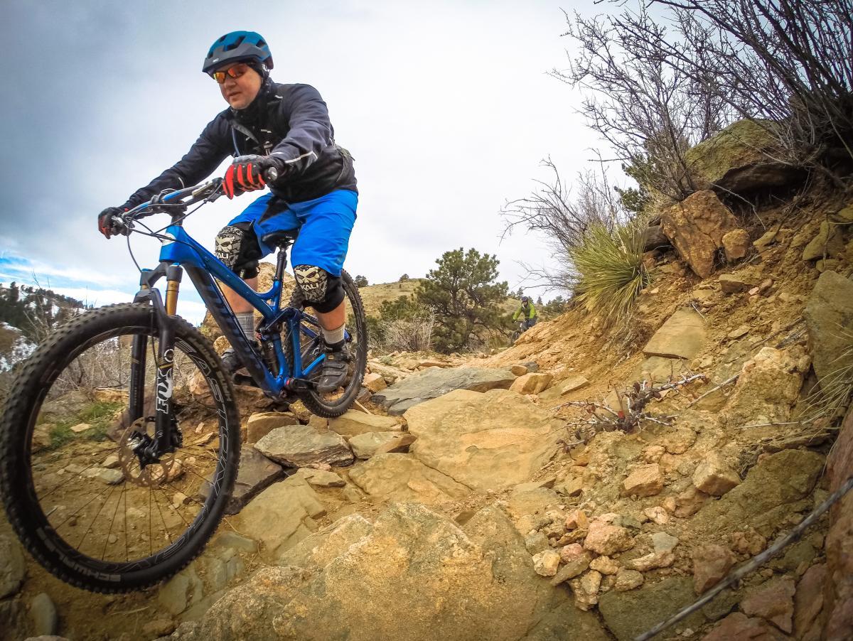 Mountain biker maneuvering over rocky terrain on a trail, wearing a helmet and protective gear, with a scenic backdrop of hills and sparse vegetation under a cloudy sky. Apex Park mountain bike trail.